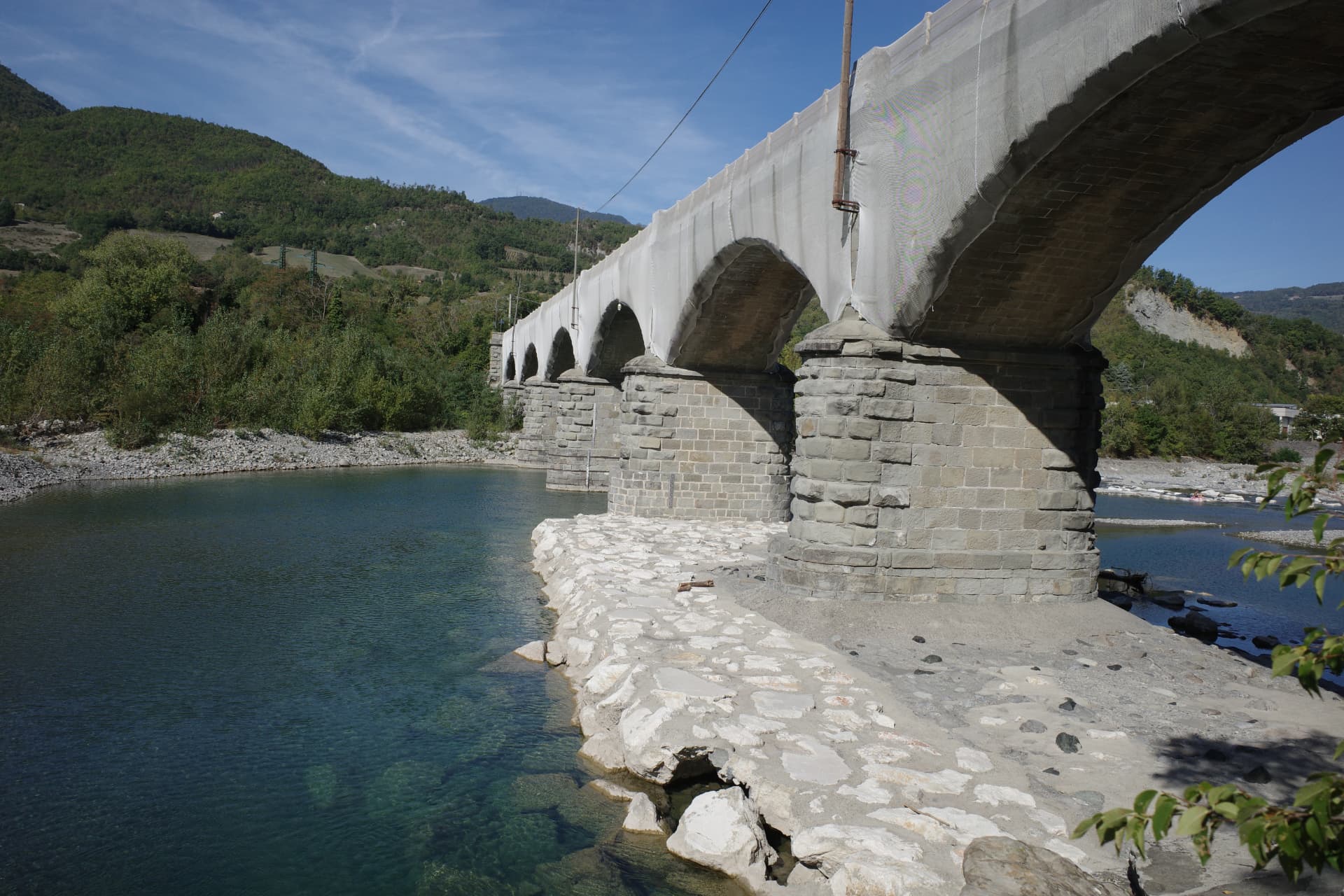 Stenpelare och valv under Ponte Gobbo, delvis täckta av nät, vid klart grönblått vatten