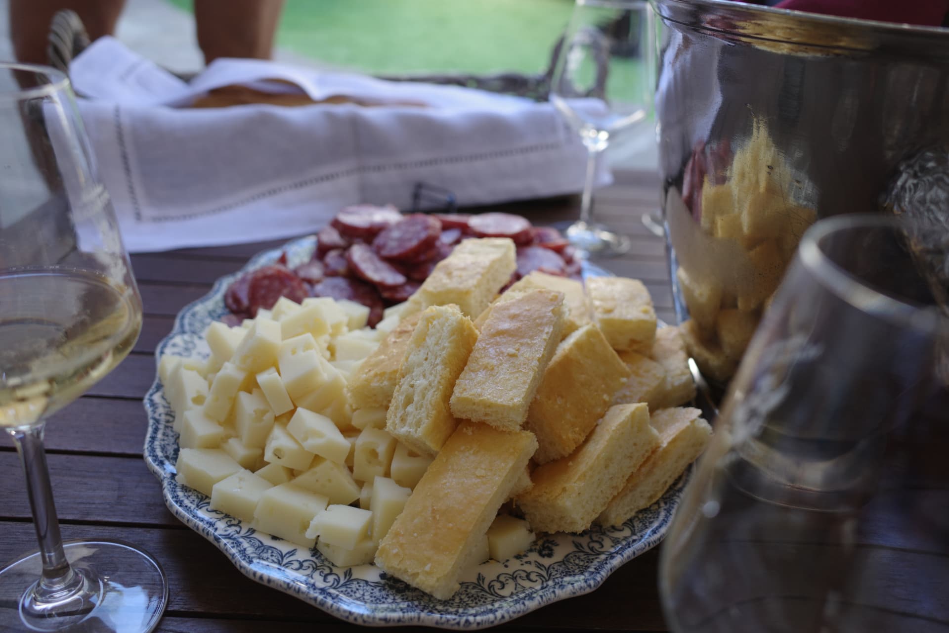 Fat med ost, bröd och salami på ett mörkt träbord, flankerat av ett glas vitt vin och en vinkylare